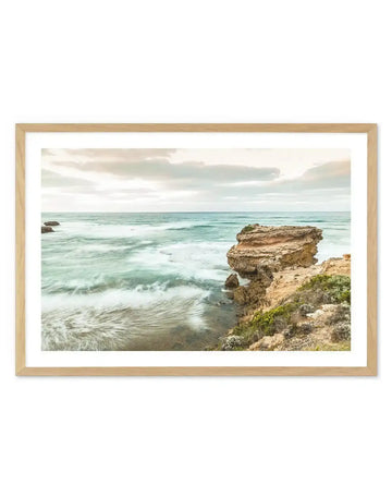 Framed photography print of a coastal scene with a large rock formation on the right and waves crashing on the shore, displayed in a natural oak frame with white border. The sky is overcast with soft grey and white clouds, and the ocean is a mix of turquoise and deep blue, with long exposure creating a smooth, misty effect on the water.