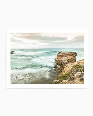 Framed photograph of a coastal scene with a large rock formation on the right, extending into the ocean. The sky is overcast with soft, muted clouds. The ocean water is a blend of turquoise and teal, with long exposure creating a smooth, ethereal effect on the waves as they break on the shore. The foreground features a rocky coastline with sparse, dry green and brown vegetation. The overall mood is serene and tranquil.