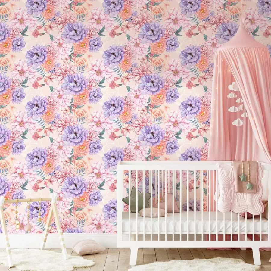 Blooming Brightly Wallpaper displayed in a nursery room featuring a dense, repeating floral pattern with purple peonies, blush pink dahlias, peachy-orange roses, and smaller pink flowers with green foliage on a cream background. The wallpaper covers the wall behind a white crib with pink fabric canopy. Watercolor-style illustration with soft, painterly textures throughout the design.