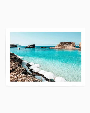 "Framed photography art print of the Blue Lagoon in Malta, featuring crystal clear turquoise water and a rocky coastline with white beach umbrellas. The composition includes several boats anchored in the lagoon and large rock formations rising from the sea under a clear blue sky, with a white frame and white border."