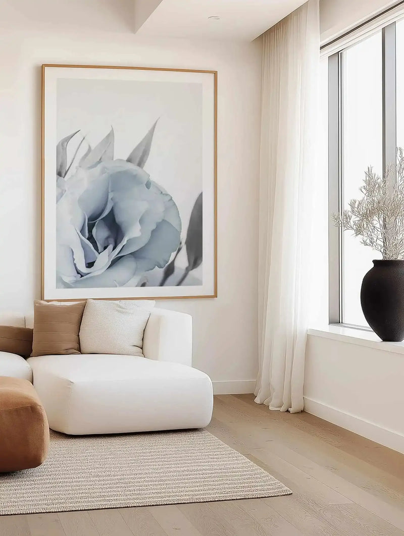Framed art print of a blue lisianthus flower depicted in soft watercolor style, featuring pale blue and gray tones with delicate petals and green leaves. The wooden frame has a natural light oak finish with a white border. Displayed above a curved white modern sofa in a bright, minimalist living room with cream curtains, light wood flooring, and a black ceramic vase on the windowsill.