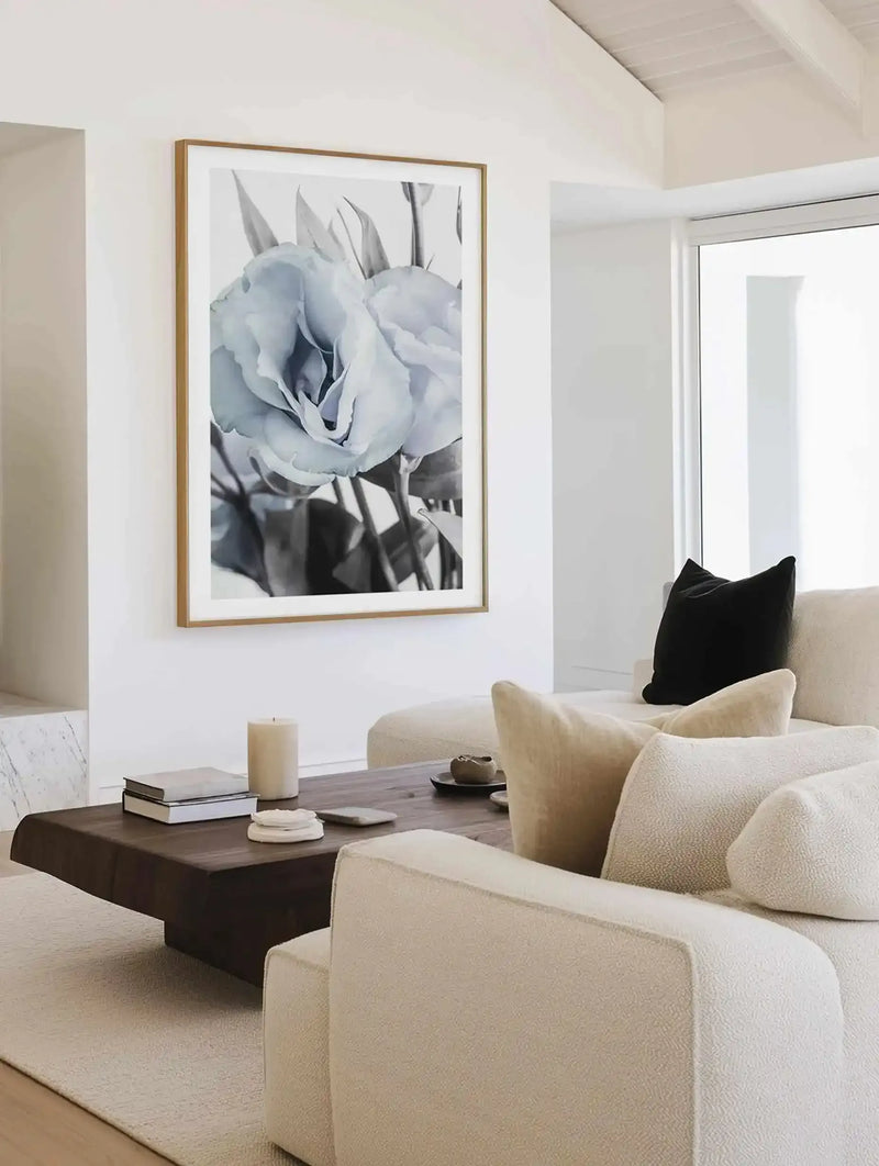 Blue Lisianthus IV art print displayed in a natural wood frame with white border, hung on a white wall above a dark wood coffee table in a minimalist living room. The print features a soft, monochromatic photograph of pale blue lisianthus flowers with delicate petals and green stems against a light background. The room includes cream-colored upholstered seating with beige pillows and a black accent pillow, a cream area rug, and natural light from large windows.