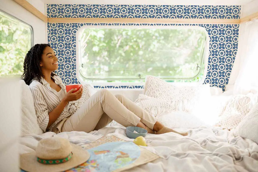 Lifestyle shot of a woman relaxing in a camper van decorated with Blue Tiling Wallpaper. The wallpaper features a repeating pattern of four-petal flowers in deep blue with a cream center, surrounded by blue leaves and set against a cream background. The woman is sitting on a bed with white bedding and pillows, holding a red mug, and looking out a window at green trees. A straw hat and a map are on the bed in the foreground.