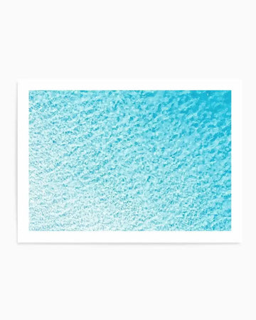 Aerial photography art print of clear turquoise ocean water with a subtle gradient from lighter aqua in the foreground to deeper blue in the distance. The water's surface is textured with small, shimmering ripples and waves, suggesting movement and light reflection. Printed on fine art paper with a matte finish.