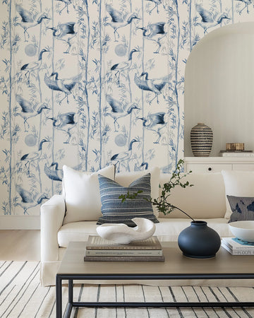Lifestyle shot of a living room featuring blue bamboo wallpaper. The wallpaper has a repeating pattern of hand-painted bamboo stalks and leaves in various shades of blue, with several cranes in different poses, also in blue and white, scattered throughout the design. The wallpaper covers the wall behind a white sofa with blue and white striped cushions. A light wood coffee table with books, a white sculptural bowl, a dark blue vase with a green branch, and a striped rug are visible in the foreground.