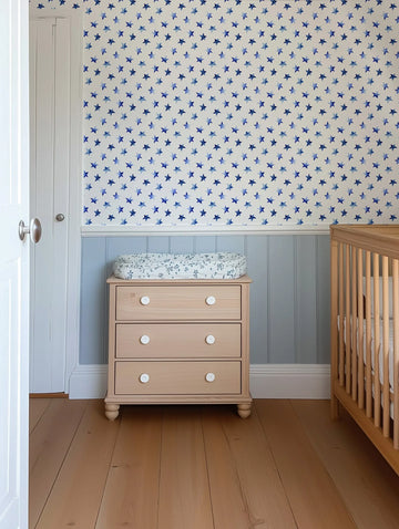 Nursery:
Blue Watercolour Stars Wallpaper installed in a baby's nursery, featuring a repeating pattern of hand-painted stars in varying shades of blue on a white background. The wallpaper covers the upper half of the wall, above a light blue wainscoting. A light wood changing table with white knobs and a floral changing pad sits against the wall, next to a matching wooden crib.