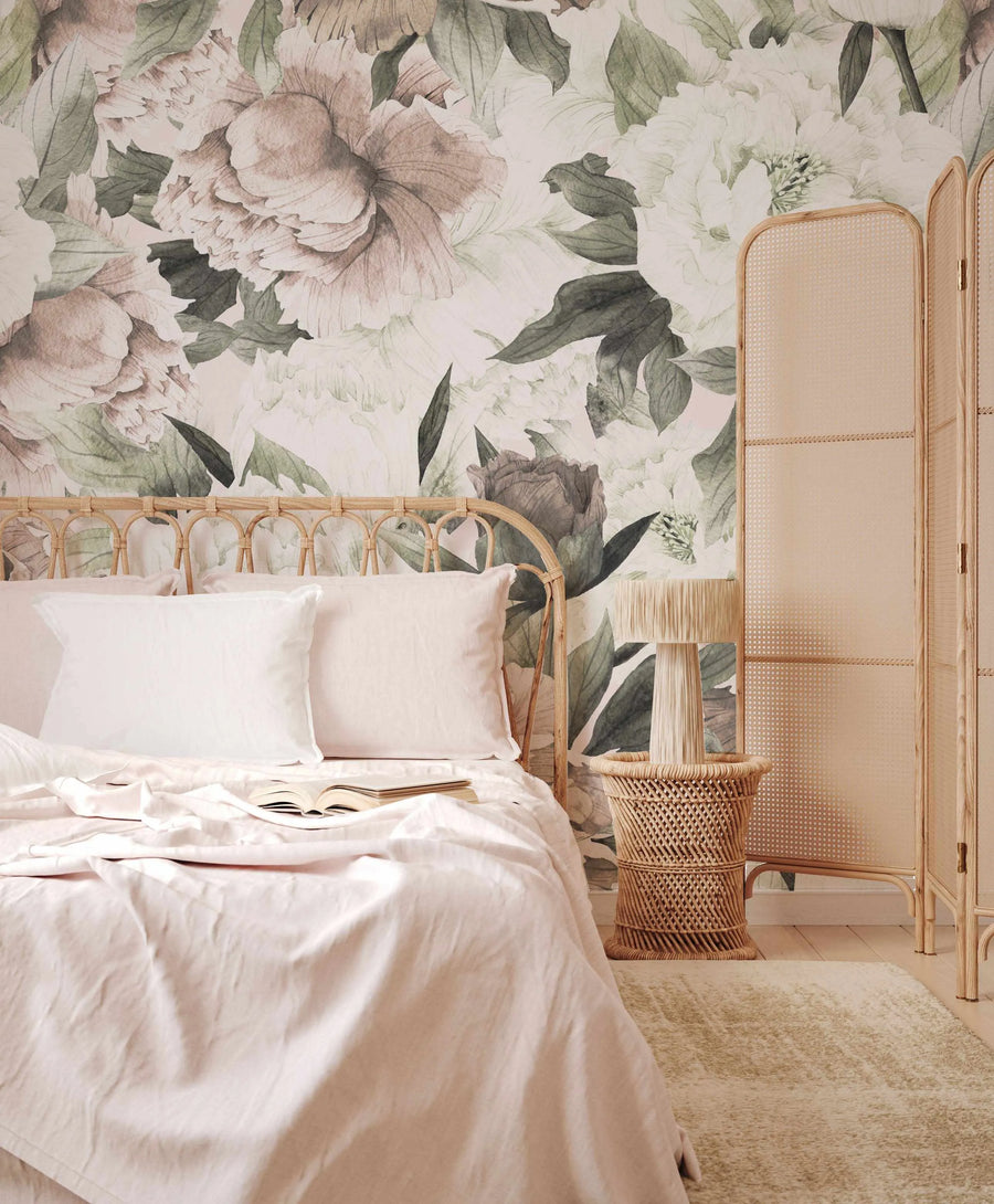 Blush Peony Floral Wallpaper Mural displayed as a feature wall in a serene bedroom. The wallpaper features large-scale, hand-painted peony blooms in soft blush pink, dusty rose, and creamy white, interspersed with muted sage green leaves, creating a delicate watercolor effect. The room includes a light wood rattan bed frame with white pillows and blush pink linen bedding, a light wood rattan side table with a lamp, a beige textured rug, and a light wood rattan room divider.