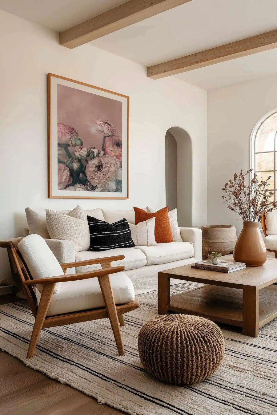 Framed art print featuring a watercolor illustration of blush pink and mauve ranunculus flowers with green stems and foliage against a soft dusty rose background, displayed in a natural wood frame with white border on a white wall above a cream linen sofa with decorative pillows in a minimalist living room.