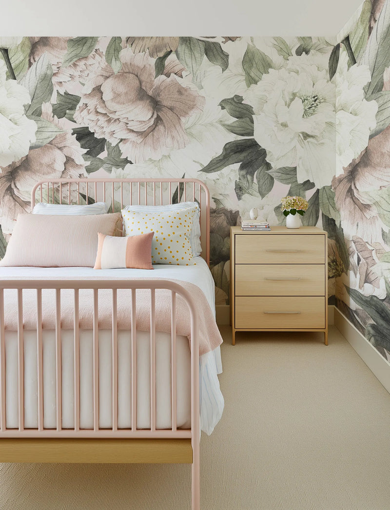 "Blush Peony Floral Wallpaper Mural displayed in a child's bedroom with a light pink metal bed frame and light wood nightstand. The wallpaper features large-scale, hand-painted peony blooms in shades of blush pink, cream, and muted sage green, creating a soft, romantic atmosphere. The bed is dressed with light pink and white bedding, and the nightstand holds a small white vase with flowers and a stack of books. The room has a light beige carpet."