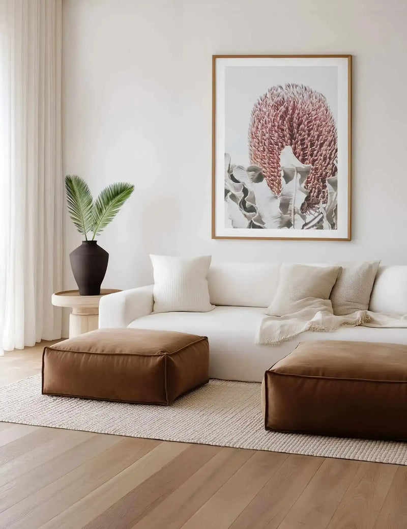 Lifestyle shot of a minimalist living room featuring a large framed art print above a white sofa. The print is a close-up photograph of a Blushing Banksia flower with a large, textured pink bloom and muted green leaves, displayed in a natural oak frame with a white border. The room includes two brown leather ottomans on a textured cream rug, and a dark brown vase with green palm fronds on a light wood side table.