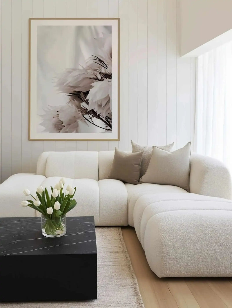 Large framed art print displayed above a curved white sofa in a minimalist living room. The print features a close-up black and white photograph of several "blushing bride" protea flowers with delicate, feathery petals, with natural wood frame and white border. The room includes a black marble coffee table with a vase of white tulips, and a textured cream rug on a light wood floor.