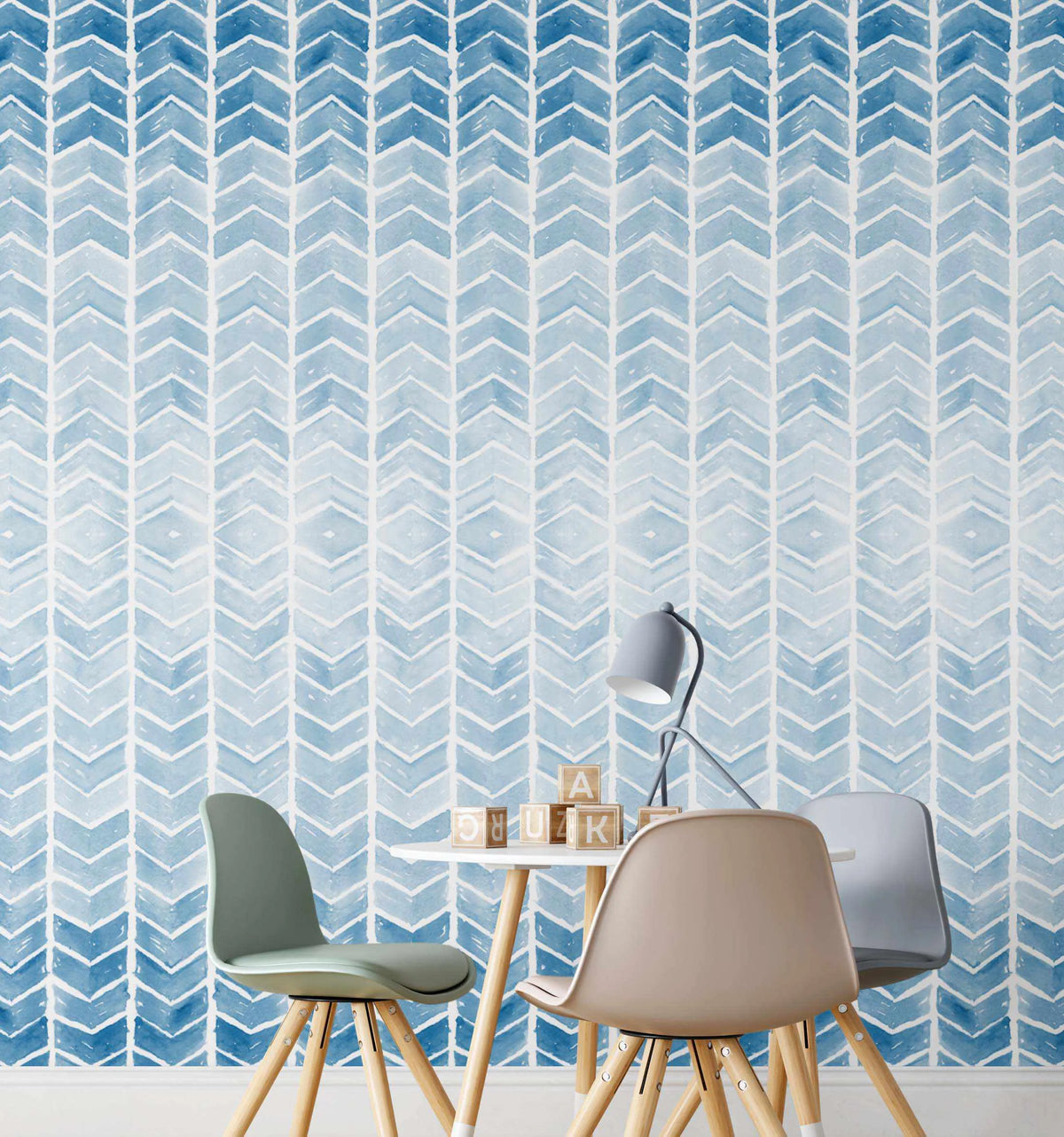 Peel and stick wallpaper featuring a repeating pattern of hand-painted chevron arrows in varying shades of blue, from deep indigo to pale sky blue, on a soft white background. The watercolour design has a loose, organic quality with visible brushstroke texture, creating a subtle ombre effect within each arrow. The wallpaper is shown in a child's room with a small white table, three chairs in sage green, beige, and grey, and wooden alphabet blocks.