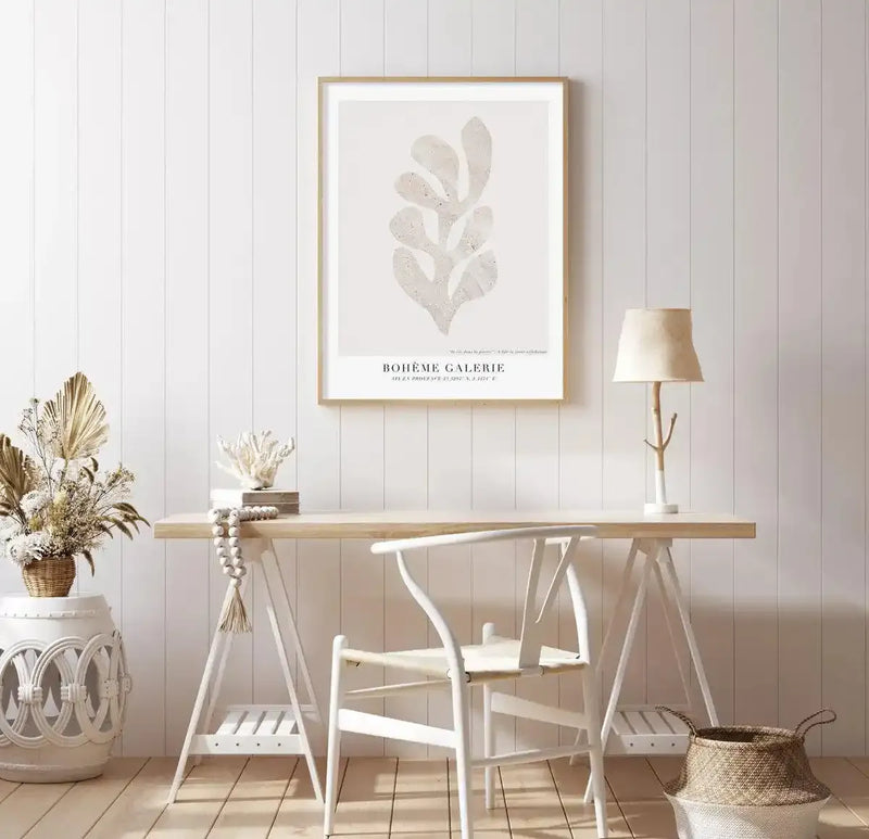 Lifestyle shot of a home office featuring the Boheme Galerie IV Art Print displayed above a light wood desk. The print features a large, abstract botanical shape in a textured, light beige against a warm cream background, framed in natural oak with a white border. The room has white vertical paneling, a white wishbone chair, a white ceramic side table with dried floral arrangements, and a small table lamp with a natural branch base.