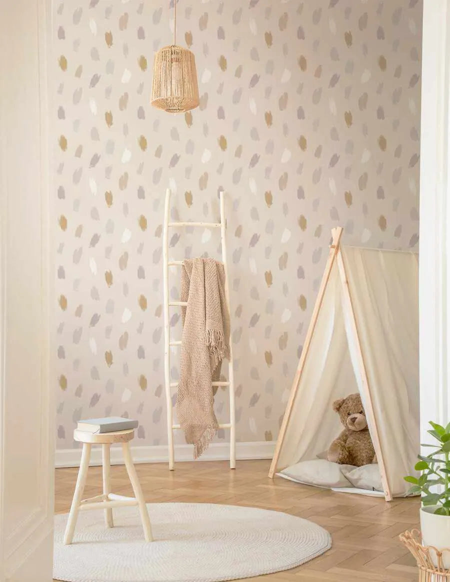 Lifestyle shot of a child's room featuring Boheme Paint Spots Wallpaper. The wallpaper has a repeating pattern of irregular, hand-painted brushstrokes in muted tones of pale gold, soft grey, and creamy white on a light beige background. The room includes a white wooden ladder with a beige knitted throw, a small white stool with books, a white round rug, and a light beige teepee tent with a teddy bear inside.