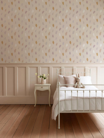 Lifestyle shot of a child's bedroom featuring Boheme Paint Spots Wallpaper on the upper wall, above a pale pink wainscoting. The wallpaper has a repeating pattern of irregular, hand-painted brushstrokes in muted shades of dusty rose, pale lavender, warm beige, and soft white on a light cream background. A white metal bed with a white duvet and a pink throw pillow sits to the right, with a small white bedside table and a vase of white flowers to the left. The room has light wooden floorboards.