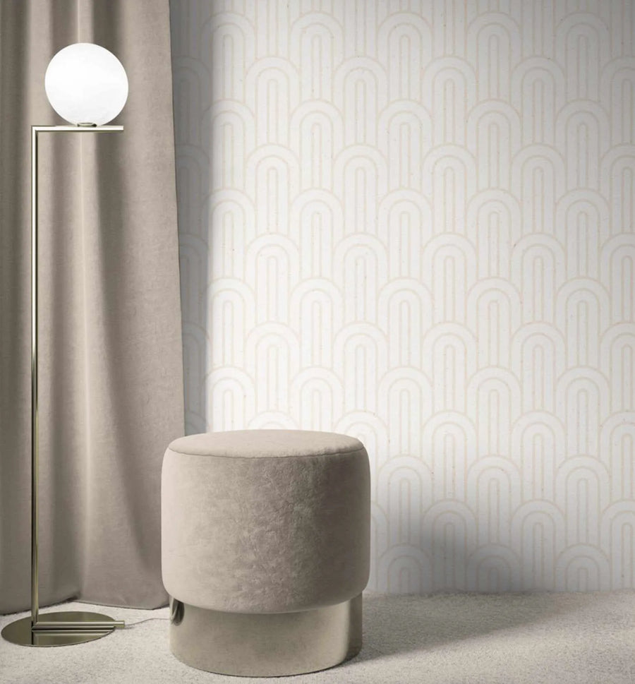 Lifestyle shot of a room featuring Bohemian Arches | Almond Wallpaper. The wallpaper has a repeating pattern of light almond-coloured arches on a soft white background, creating a subtle, textured look. A tall, modern floor lamp with a spherical white light is positioned to the left, next to a flowing, light taupe curtain. A round, velvet ottoman in a similar taupe shade sits on a light grey carpet in the foreground.