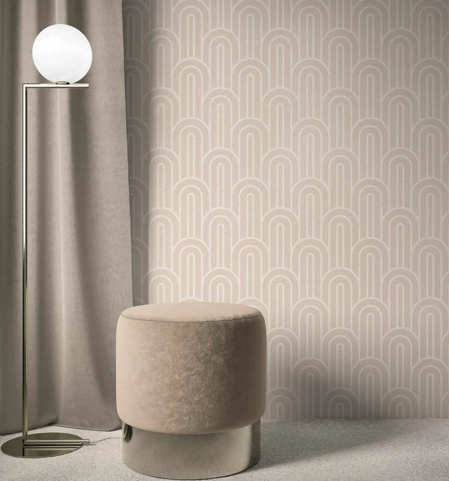 Lifestyle shot of a room with "Bohemian Arches | Latte" wallpaper. The wallpaper features a repeating pattern of rounded arches in a lighter latte shade against a slightly darker latte background, creating a subtle, textured look. A modern floor lamp with a spherical white globe light and a brass-toned stand is positioned on the left, next to a taupe-colored curtain. A round, upholstered stool in a similar taupe hue with a metallic base sits in the foreground on a light grey carpet.
