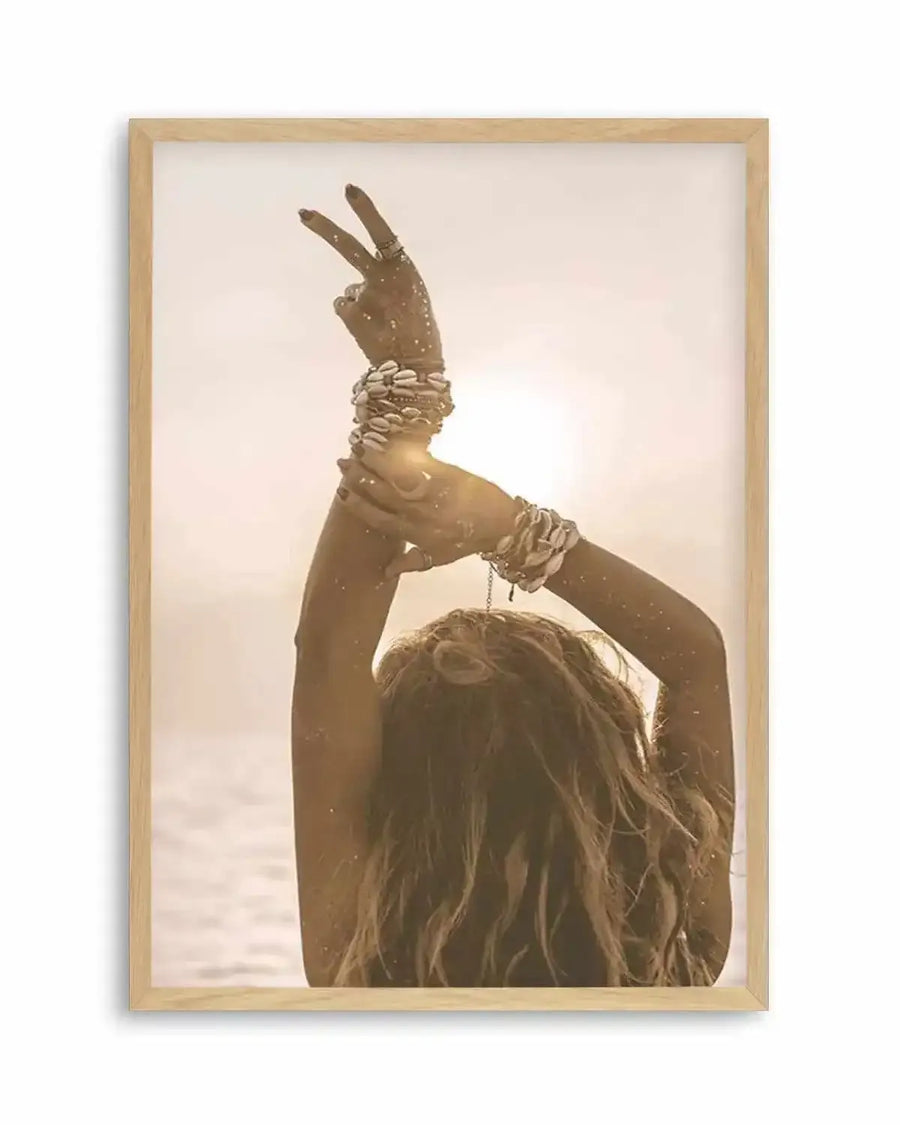 Framed photography art print featuring a bohemian girl with her back to the viewer, arms raised in a peace sign against a bright, sun-drenched background. She has long, wavy brown hair and is adorned with multiple shell bracelets on each wrist. The image has a warm, golden hue, suggesting a sunset or sunrise over water, and is displayed in a natural oak frame with a white border.