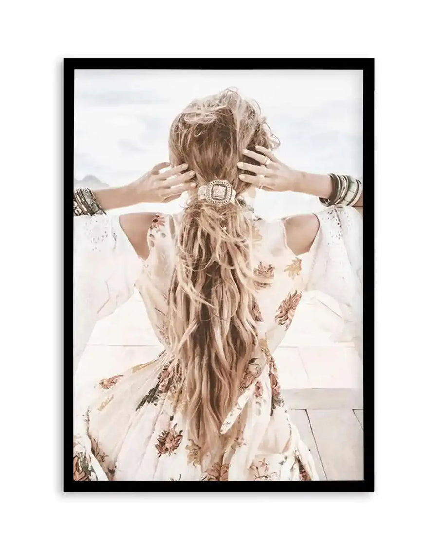 Framed photography art print of a woman from behind, with long blonde hair tied in a low ponytail with an ornate silver hair tie, wearing a white floral dress with wide sleeves, and multiple bangles on each wrist. The woman is looking out at a bright, hazy outdoor scene, with her hands resting on her head. The print is displayed in a black frame with a white border.