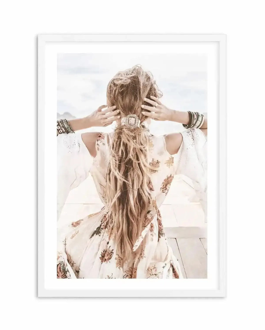 Framed photography art print featuring a woman from behind, with long wavy blonde hair tied in a low ponytail with an ornate hair tie, wearing a white bohemian-style dress with floral patterns in muted orange and brown tones, and multiple bracelets on both wrists. The woman is looking out towards a bright, slightly out-of-focus background of what appears to be a beach or open landscape, with a white frame and white border.