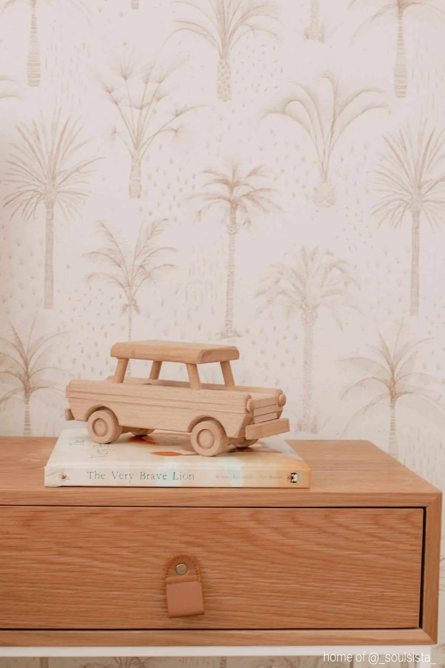Bohemian Palms wallpaper featuring a repeating pattern of hand-drawn palm trees in a soft sepia tone on a light cream background. The wallpaper is visible behind a light wood bedside table with a single drawer and a tan leather pull. On the table, a wooden toy car sits atop a book titled "The Very Brave Lion".
