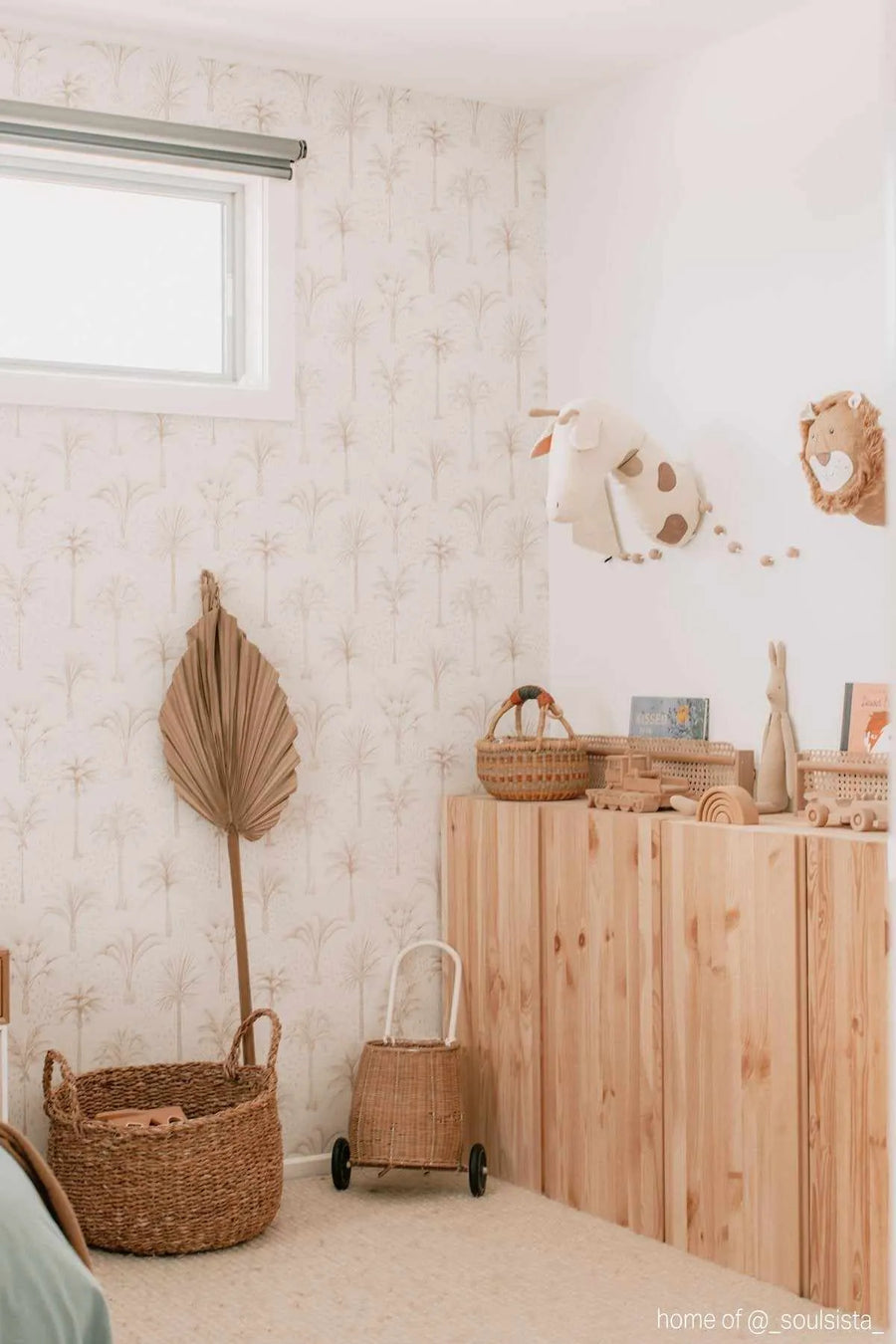 Lifestyle shot of a nursery featuring Bohemian Palms Wallpaper. The wallpaper has a repeating pattern of hand-drawn palm trees in a soft, muted beige and cream palette, creating a subtle tropical feel. The room includes a window with a grey blind, a light wood cabinet with various wooden toys and woven baskets, a large dried palm frond, and a stuffed animal head mounted on the wall.