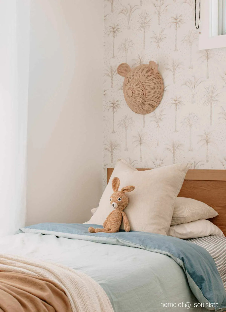 Lifestyle shot of a child's bedroom featuring Bohemian Palms Wallpaper. The wallpaper has a repeating pattern of hand-drawn palm trees in a soft beige-grey on a light cream background, creating a serene, natural feel. The wallpaper covers the wall behind a wooden bed with light blue and white bedding, and a beige knitted bunny toy sitting on a cream pillow. A woven wicker bear head wall hanging is also visible on the wallpapered wall.