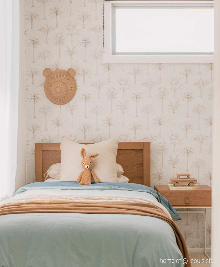 "Bohemian Palms peel and stick wallpaper featuring a repeating pattern of hand-drawn palm trees in muted beige and cream on a soft white background, displayed in a child's bedroom. The wallpaper covers the wall behind a single bed with a light wood headboard, light blue duvet, and a brown knitted bunny toy. A round woven bear-ear wall hanging is visible on the left, and a small wooden bedside table with a toy car is on the right."