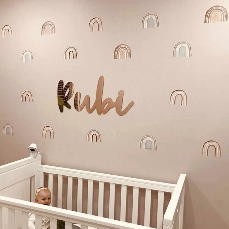 Nursery:
"A baby girl in a white crib looks up at a wall decorated with Bohemian Rainbows wall decals. The decals feature repeating watercolour-style rainbows in muted tones of dusty rose, pale grey, and warm cream, scattered across a light pink wall. A reflective gold name decal spelling 'Rubi' is centered above the crib."