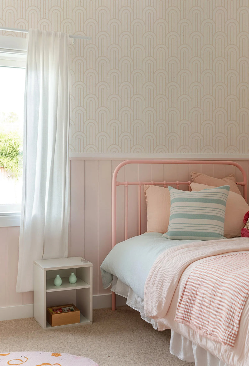 Lifestyle shot of a child's bedroom featuring a wallpaper with a repeating pattern of bohemian arches in a light almond or beige tone on a cream background. The wallpaper is displayed above a pale pink wainscoting. The room includes a pink metal bed frame with light blue and pink bedding, a striped blue and white pillow, and a white bedside table with light green vases.