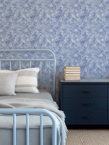 Lifestyle shot of a bedroom featuring a light blue metal bed frame with a grey duvet and throw, and two pillows (one white with grey stripes, one white with a black grid pattern). To the right, a dark blue three-drawer nightstand with black handles holds a stack of books. The wall behind the bed is covered in a denim blue and white chevron pattern wallpaper with a textured, distressed look. The floor is covered with a light brown woven rug.