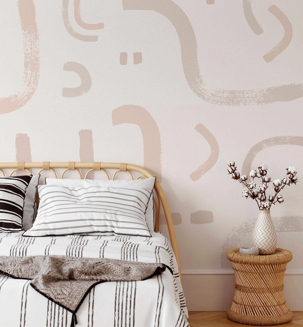 Lifestyle shot of a bedroom featuring a peel and stick wallpaper mural with an abstract pattern of organic brushstrokes in varying shades of dusty rose, beige, and light brown on a soft white background. The wallpaper is displayed behind a light wood rattan bed with white and black striped bedding and a textured grey throw. A woven rattan side table with a vase of cotton branches sits to the right of the bed.