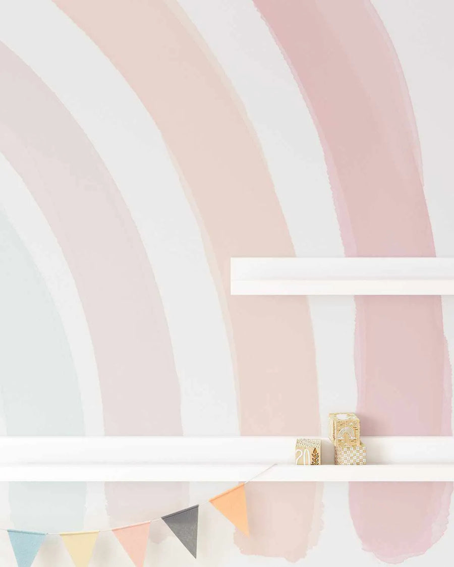"Peel and stick wallpaper featuring a large-scale, hand-painted rainbow mural with wide, arcing stripes in soft pastel shades of dusty rose, blush pink, and pale blue on a white background. The design has a loose, organic watercolour quality with visible brushstroke texture. A white floating shelf is visible on the right, holding two small decorative wooden blocks, and a string of pastel-coloured bunting hangs below."