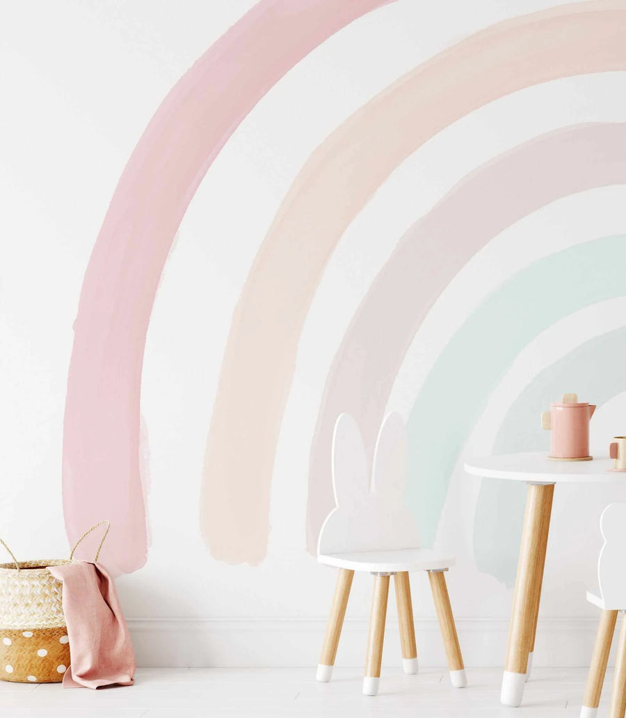 Kids Room:
Boho Rainbow Wallpaper Mural displayed on a white wall in a child's room, featuring a large-scale, hand-painted rainbow design with five arched stripes in soft, muted pastel colours: dusty rose, pale peach, light beige, soft pink, and mint green. The wallpaper has a watercolour texture with visible brushstrokes. A woven basket with a pink linen cloth sits on the floor to the left, and a white round table with two white bunny-ear chairs and a pink toy teapot are visible on the right.