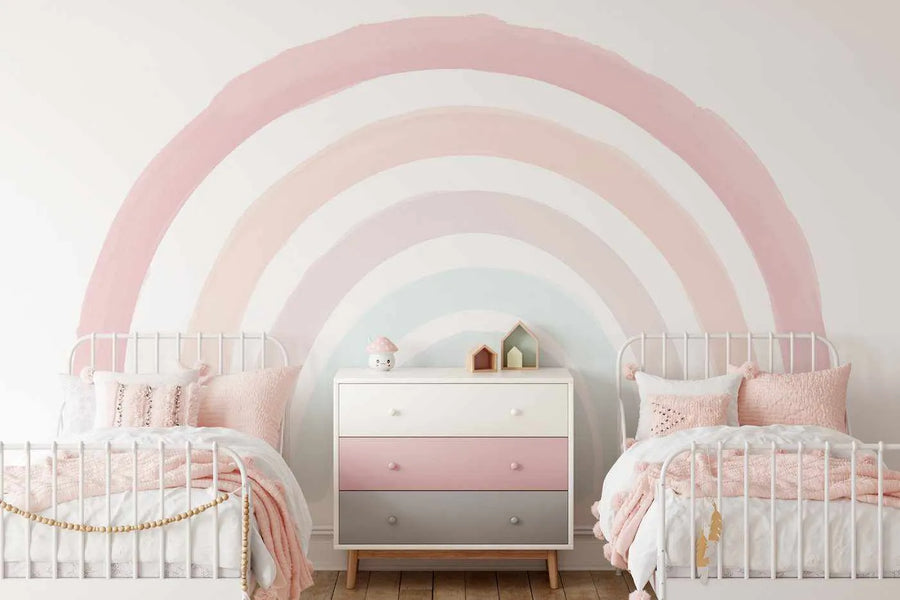 Two white metal-frame twin beds with pink bedding are positioned on either side of a white and pink dresser, all against a white wall featuring a large, hand-painted style rainbow mural. The rainbow has wide, arcing bands in shades of dusty rose, pale peach, soft lavender, and light blue, creating a whimsical and serene atmosphere in the children's bedroom.
