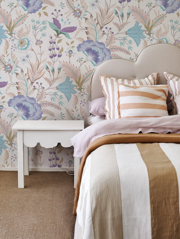 Boho Blooms wallpaper featuring a repeating pattern of large-scale floral and botanical motifs in shades of lavender, periwinkle, teal, blush pink, and sage green on a cream background, displayed in a bedroom above a bed with a curved, upholstered headboard. The bed is dressed with a light pink duvet, a brown throw, and striped pillows in white and light brown. A white scalloped bedside table is visible on the left.