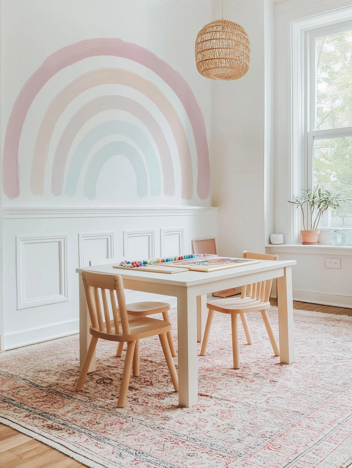 Kids Room:
Boho Rainbow Wallpaper Mural displayed on a white wall above white wainscoting in a bright kids' play area. The mural features a large, hand-painted rainbow design with soft, muted arches in shades of dusty rose, pale peach, light grey, and soft teal, giving a watercolour effect. A light wood children's table and chairs are positioned in front of the mural on a patterned rug, with a woven pendant light hanging from the ceiling and a window with a potted plant on the sill to the right.