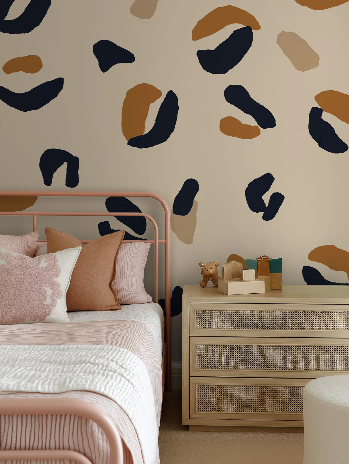 Lifestyle shot of a bedroom featuring a peel and stick wallpaper mural with a bold leopard print pattern in abstract shapes of deep navy blue, warm caramel brown, and light beige on a soft cream background. The wallpaper covers the wall above a pink metal bed frame with a white and pink striped duvet and various pillows in shades of pink and light brown. To the right of the bed is a light wood dresser with woven cane drawer fronts, topped with wooden blocks and a small brown teddy bear figurine.
