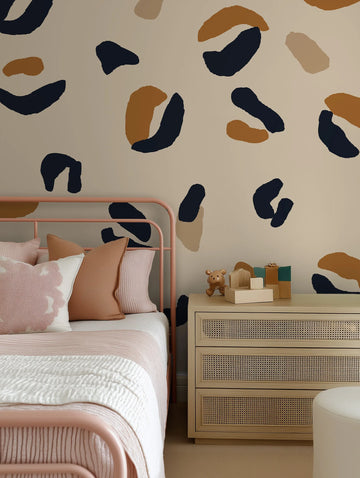 Lifestyle shot of a bedroom featuring a peel and stick wallpaper mural with a bold leopard print pattern in abstract shapes of deep navy blue, warm caramel brown, and light beige on a soft cream background. The wallpaper covers the wall above a pink metal bed frame with a white and pink striped duvet and various pillows in shades of pink and light brown. To the right of the bed is a light wood dresser with woven cane drawer fronts, topped with wooden blocks and a small brown teddy bear figurine.