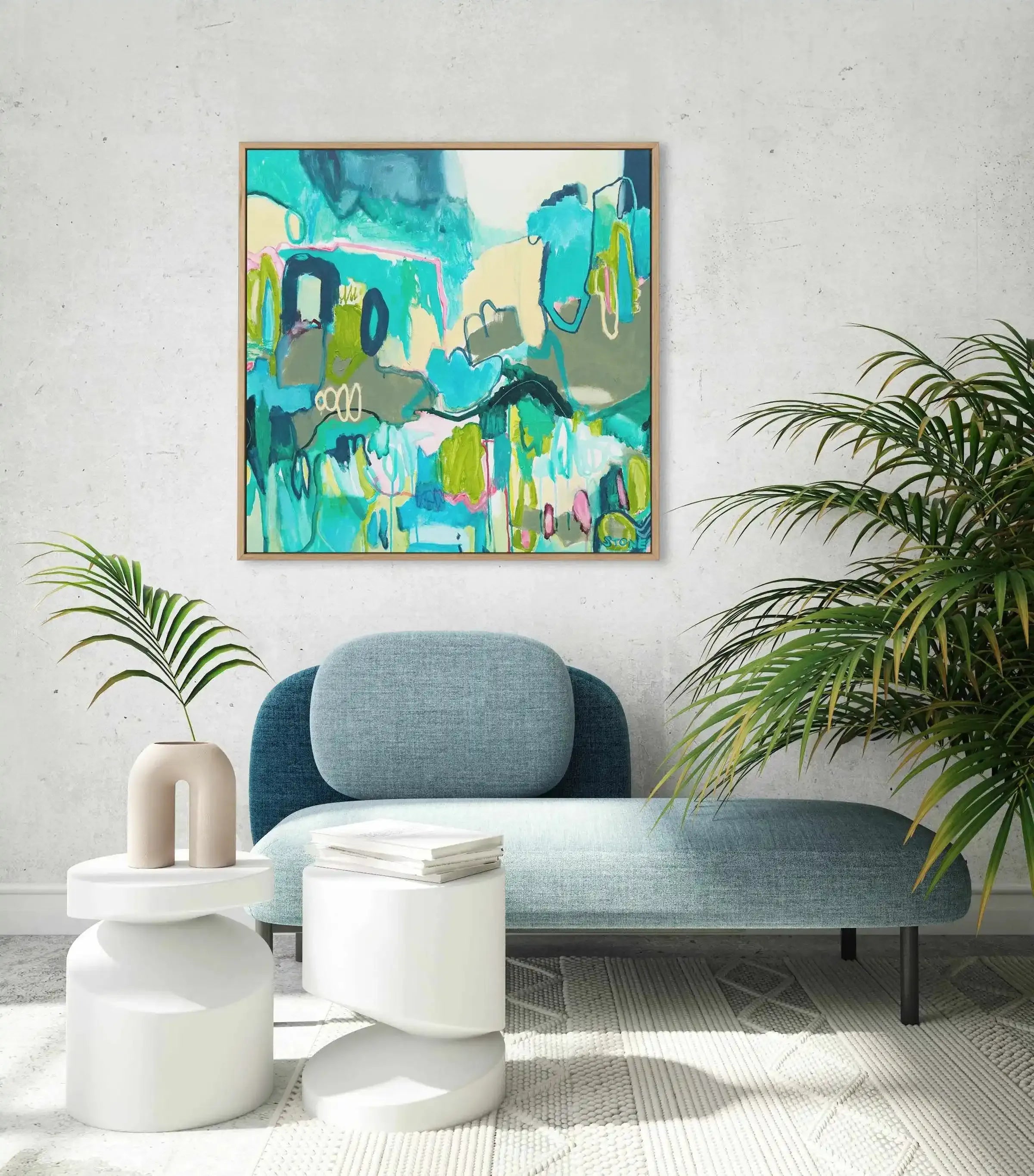 Lifestyle shot of a minimalist living room featuring a framed abstract canvas print above a blue chaise lounge. The print, titled "Bondi Blues," features an abstract composition of organic shapes and expressive brushstrokes in shades of teal, aqua, lime green, pale yellow, and touches of pink and dark blue, with a natural wood frame. The room includes two white cylindrical side tables, a beige arched vase with a single palm leaf, and a large green plant on the right.