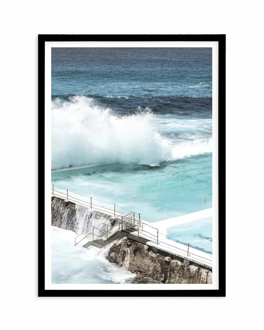 Framed art print featuring a photograph of Bondi Icebergs Pool. The image shows a dramatic seascape with turquoise swimming pool in the foreground, white metal railings along a concrete platform at the pool's edge, and rocky cliff face below. Large white-capped waves crash in the background against darker ocean waters. The composition captures the contrast between the calm, bright turquoise pool and the powerful, foamy waves beyond. Black wooden frame with white border surrounding the photograph.