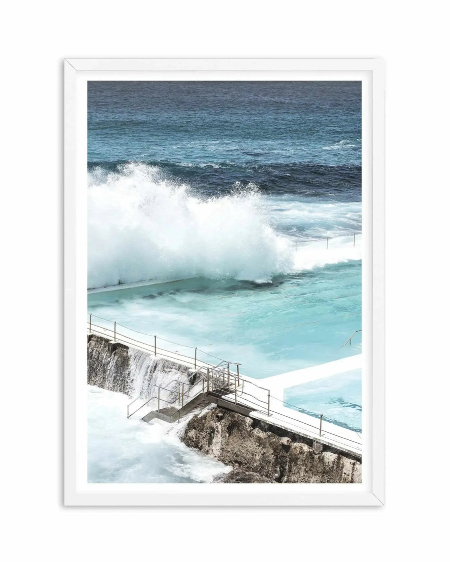 Framed photography art print of the Bondi Icebergs Pool with a white frame and white border. The image captures a large wave crashing over the edge of the ocean pool, creating a dramatic spray of white foam against the turquoise water. The pool's concrete edge and metal railings are visible in the foreground, with water cascading over the lower rock wall. The ocean beyond is a deep blue-green.