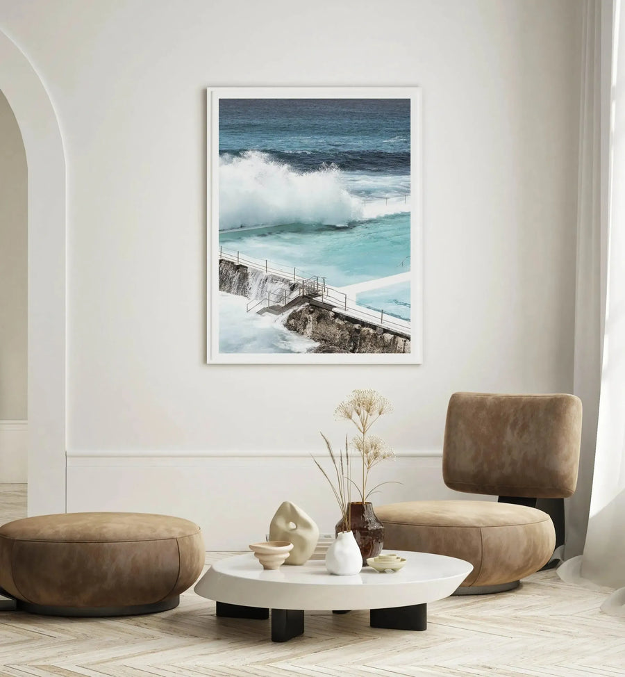 Framed photograph of Bondi Icebergs Pool featuring turquoise water with white-capped waves, a metal staircase descending a rocky cliff, and deep blue ocean beyond. White frame with white border. Displayed above a round white table with black legs in a minimalist living room with tan leather poufs and neutral décor.