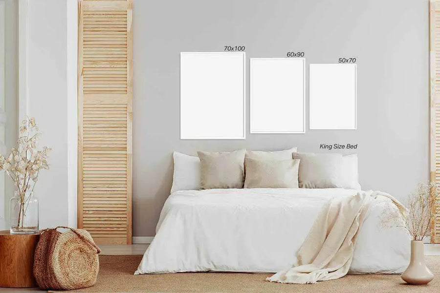 Lifestyle shot of a minimalist bedroom featuring a King Size Bed with white bedding and neutral pillows. Above the bed, three framed art prints are displayed on a light grey wall. The largest print, 70x100, is on the left, followed by a 60x90 print in the middle, and a 50x70 print on the right. All frames are white with white borders. To the left of the bed, a wooden stool holds a glass vase with dried flowers, and a woven basket sits on a brown rug. A wooden louvered screen is visible on the left wall.