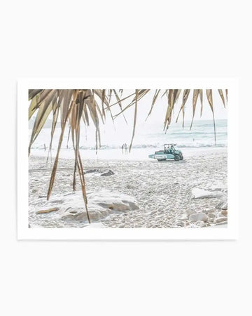 Framed photography art print of a bright coastal beach scene with natural wood frame and white border. The composition features a light blue utility vehicle with a surfboard on the back, parked on white sand near the ocean. In the foreground, dried palm fronds hang down, partially obscuring the view. Two people walk along the water's edge in the distance, under a bright, hazy sky.