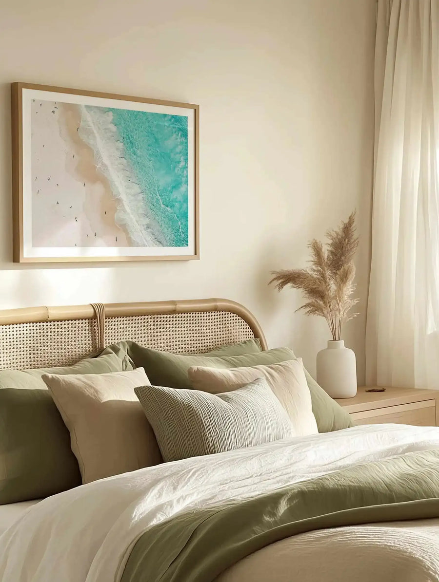Aerial photograph of a turquoise ocean with white waves and sandy beach dotted with people, displayed in a natural wood frame with white border above a rattan headbed styled with cream and sage green bedding, pampas grass in a white ceramic vase on wooden nightstand, and sheer cream curtains in background.