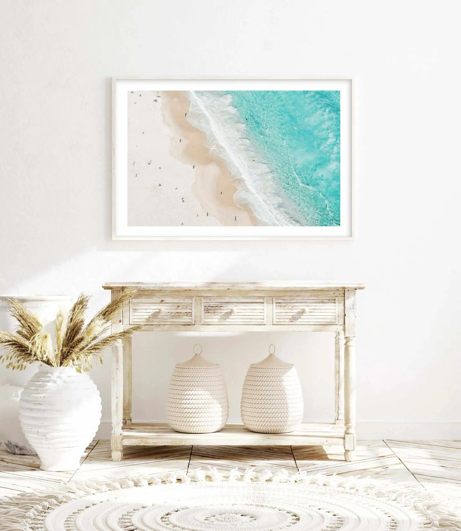 Aerial photograph of a beach coastline framed in light wood with white border, displayed above a whitewashed wooden console table. The artwork shows turquoise ocean water, white wave foam, and pale sand with scattered sunbathers viewed from above. A white ceramic vase with dried pampas grass sits to the left of the console, two cream woven baskets sit on the shelf below, and a white circular woven rug with fringe sits on the wooden floor in front.