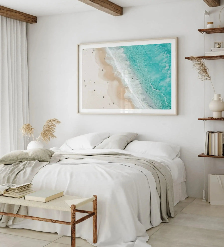 Aerial photograph framed in light wood showing a turquoise ocean with white wave foam meeting a pale sandy beach dotted with small swimmers and beach umbrellas, displayed above a bed with white linens in a minimalist coastal bedroom with wooden ceiling beams, white walls, and wooden floating shelves.
