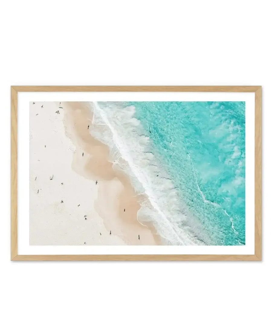 Framed aerial photograph of Bondi Beach with turquoise water, white sand, and small figures of people, displayed in a natural oak frame with white border. The composition captures the clear blue-green ocean meeting the shore with white foamy waves, and the light-coloured sand stretching back, dotted with beachgoers.