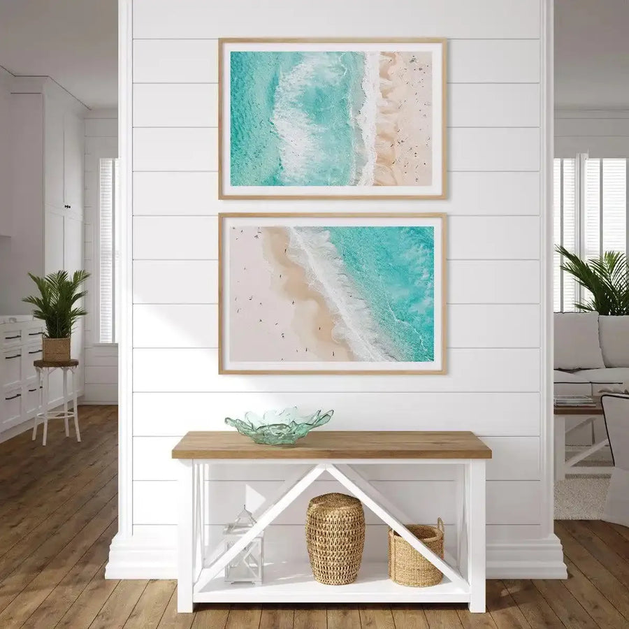 Two framed aerial photography art prints displayed vertically on a white shiplap wall in a bright, coastal-inspired entryway. Each print features a vibrant turquoise ocean meeting a sandy beach with small waves breaking on the shore, and tiny figures of people scattered along the sand. The prints are framed in natural wood with white borders. Below the prints is a white console table with a wooden top, holding a light green glass bowl, a woven basket, and a white lantern.